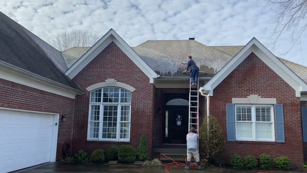 Workers using a ladder to clean the roof of a brick house with pressure washing equipment.